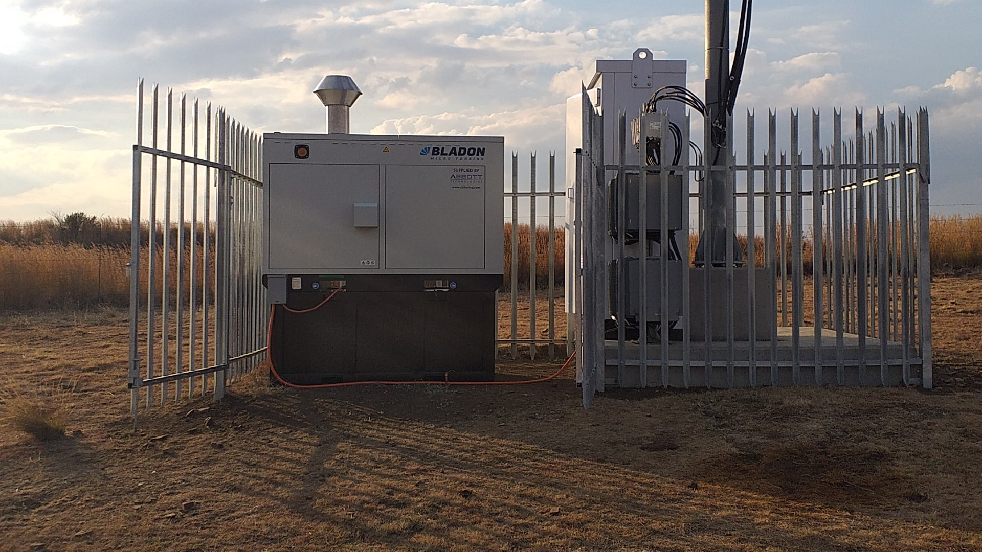 Bladon microturbine genset behind a palisade security fence at a rural off grid telecom site in West Africa, with the tower mast and dry grassland in the background.